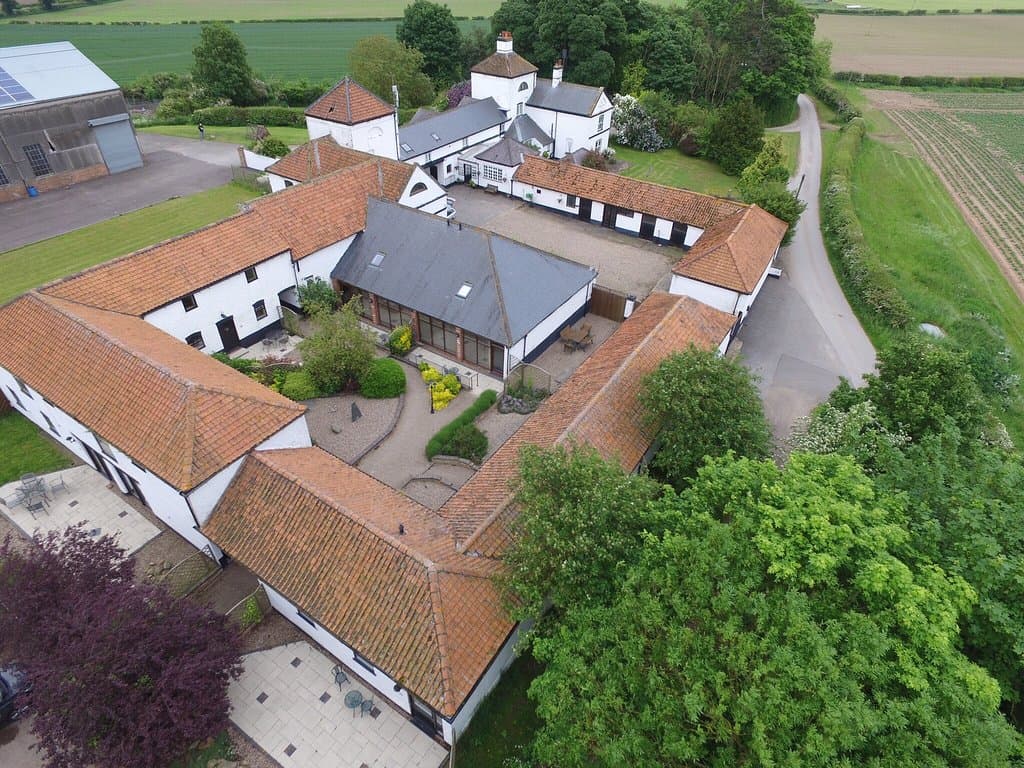 Aerial view of Blaco Hill Farm: whitewashed buildings with red pantile roofs around a courtyard garden, with fields beyond