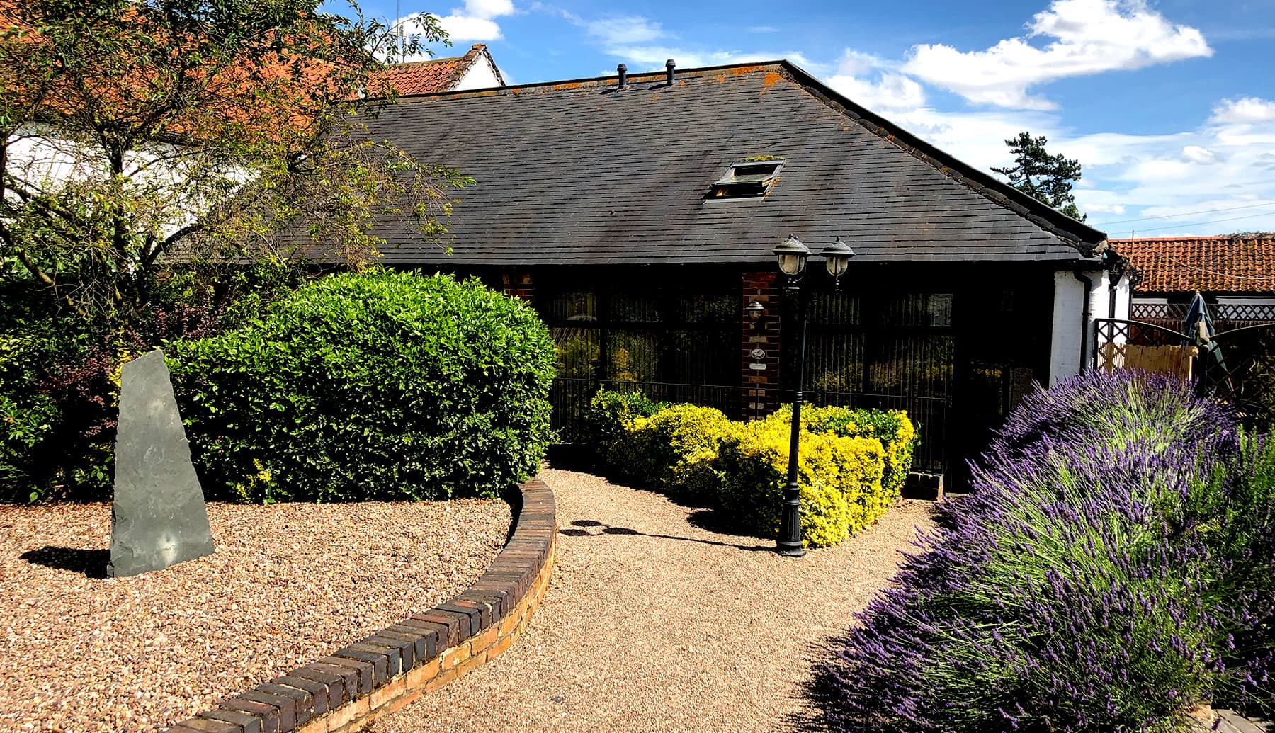 Single-storey cottage with a slate roof behind a gravel garden with lavender, shrubs, a standing stone and a lamp post