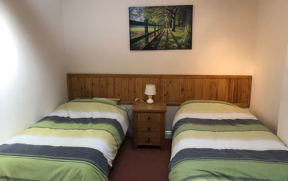 Twin bedroom with two single beds with green striped covers beneath a painting of a tree-lined path
