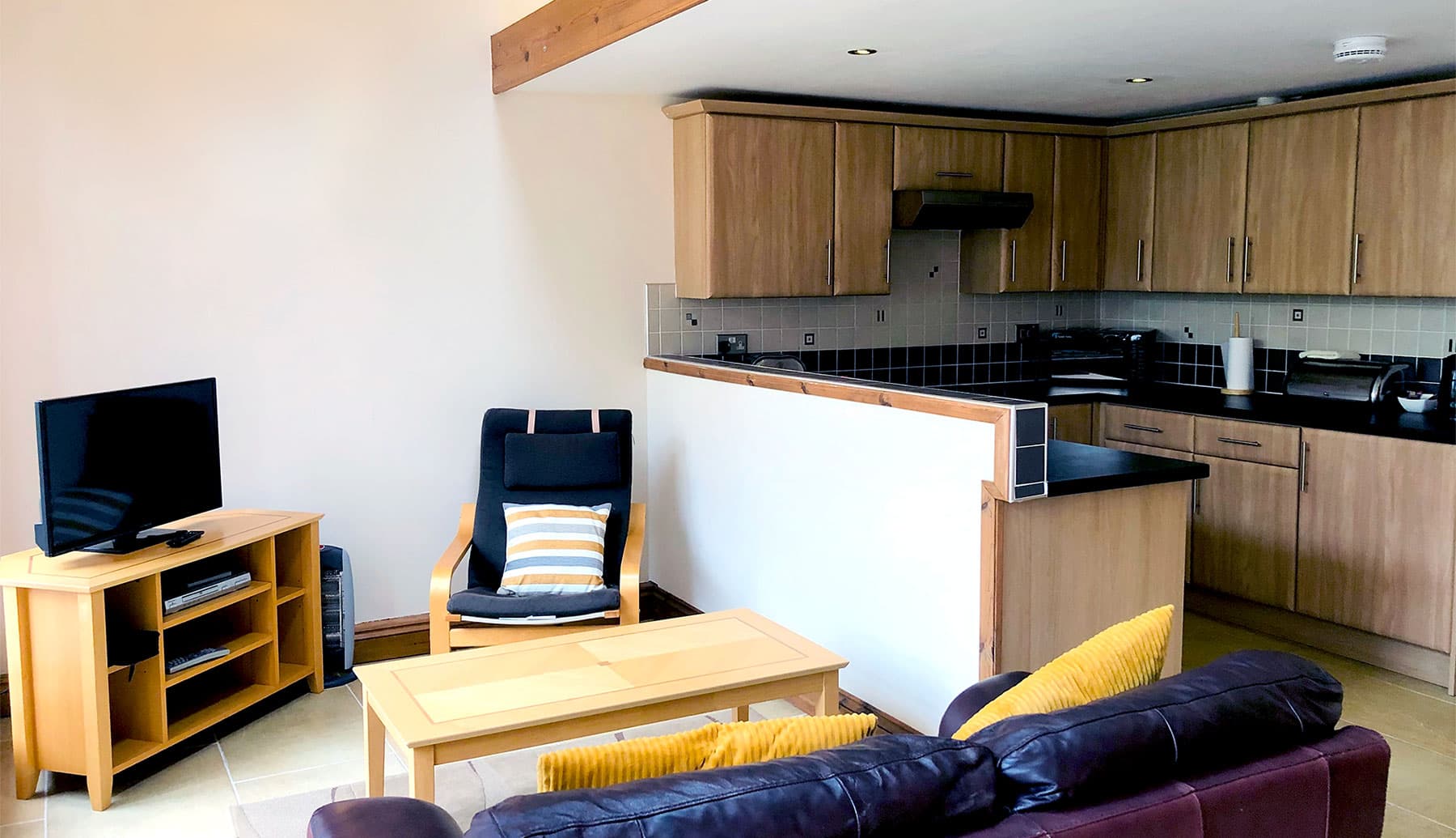 Open-plan living area with a leather sofa, armchair, coffee table and TV, and a kitchen behind a half wall