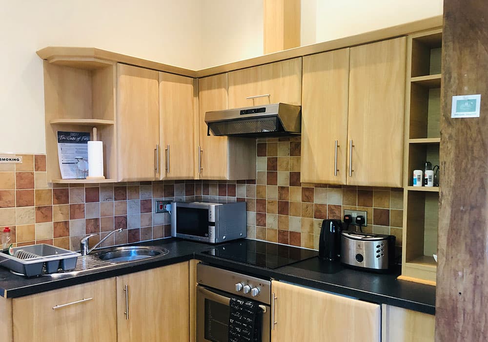 Kitchen with light oak units, a black worktop, microwave, oven and a terracotta tiled splashback