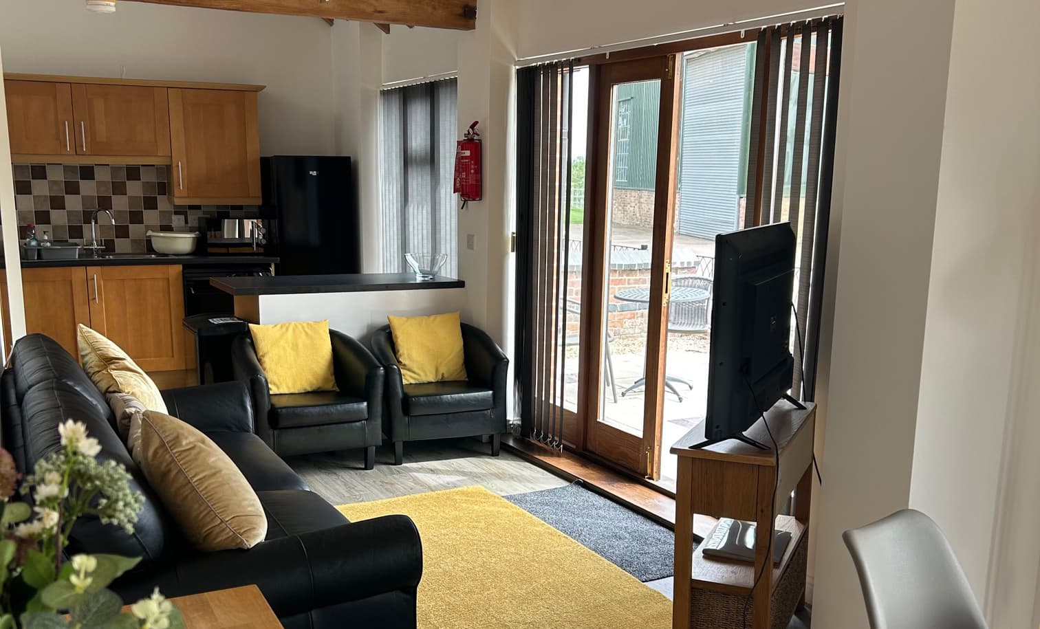 Open-plan living room with black leather sofas and armchairs, a yellow rug, a kitchen with oak units and glazed doors to the patio